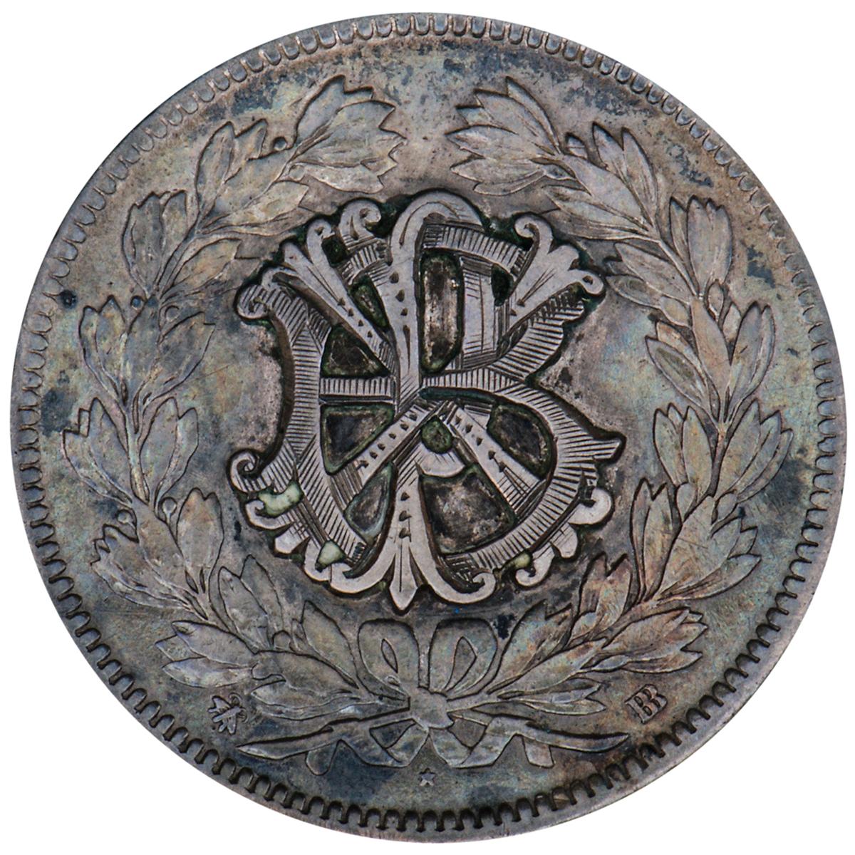 Pièce d’argent polie sur laquelle sont gravées des lettres entrelacées et empilées les unes sur les autres, entourées d’une couronne de feuilles.