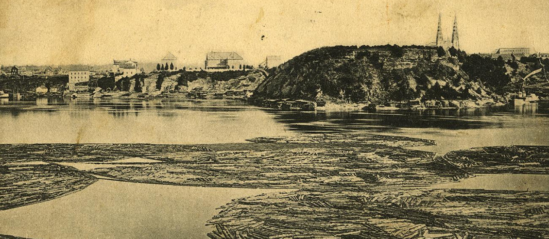 Photo en noir et blanc de billots retenus par des chaînes flottant sur une rivière devant une colline boisée.