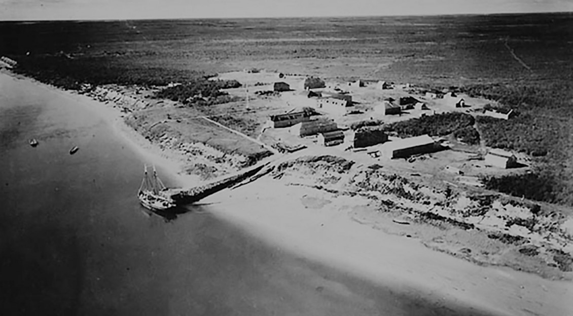 Photographie en noir et blanc montrant la vue aérienne d’une vaste terre inoccupée et d’établissements de style militaire situés près de la rive.