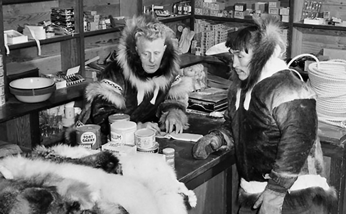 Photographie en noir et blanc montrant un homme blanc et un homme autochtone vêtus d’un parka dans une pièce remplie de provisions.