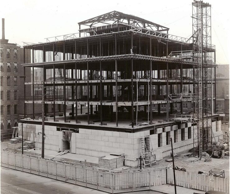 Photographie en noir et blanc de la charpente d’un immeuble carré de quelques étages dont le bas est en blocs de pierre et le reste en poutres d’acier.