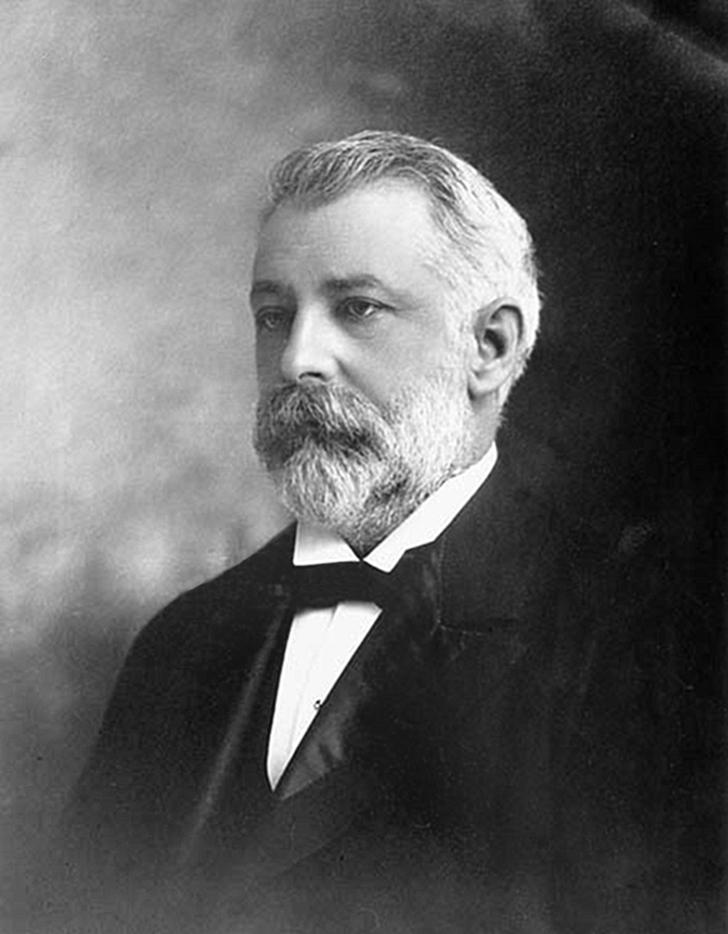 Photographie en noir et blanc d’un homme barbu aux cheveux gris portant un costume du 19e siècle.