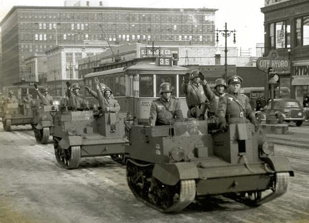 Photo en noir et blanc d’une rue de la ville, avec des véhicules de l’armée remplis d’hommes au bras droit levé en signe de salut nazi. 