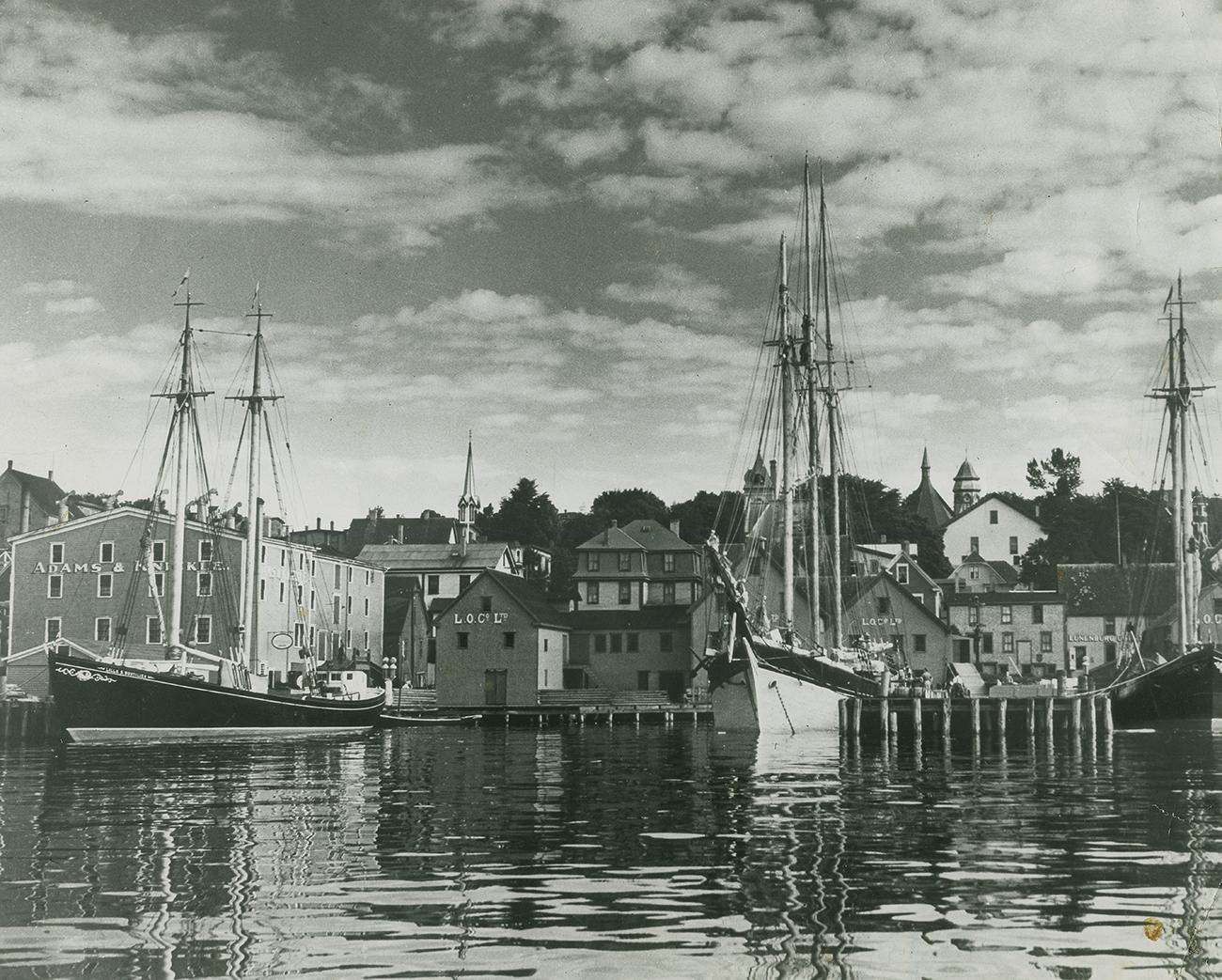 Photo en noir et blanc, bâtiments en bois au bord de l’eau, trois voiliers amarrés à un quai.