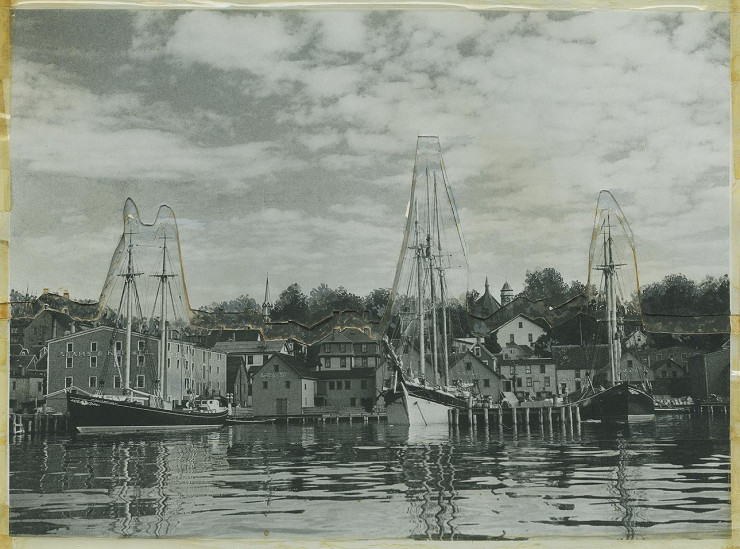 Photo en noir et blanc, bâtiments en bois au bord de l’eau, trois voiliers amarrés à un quai.