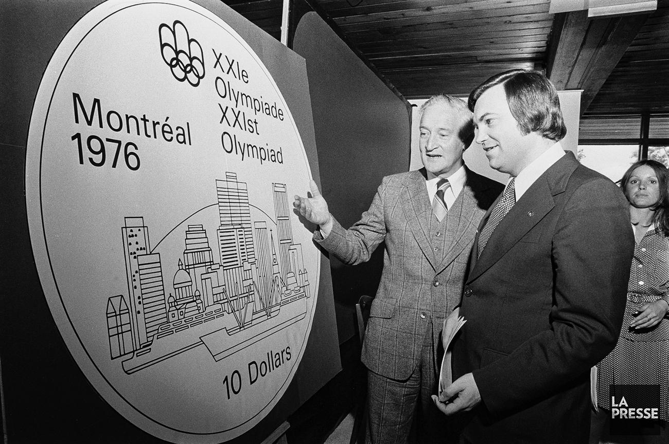 Dévoilement d’une pièce des Jeux olympiques Photo en noir et blanc de deux hommes examinant le grand dessin d’une pièce représentant une ville sous un logo olympique.