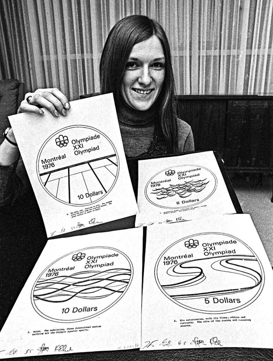 Lynda Cooper 1975 Photo en noir et blanc d’une jeune femme assise, tenant un dessin dans chaque main, avec deux autres dessins posés sur une table.