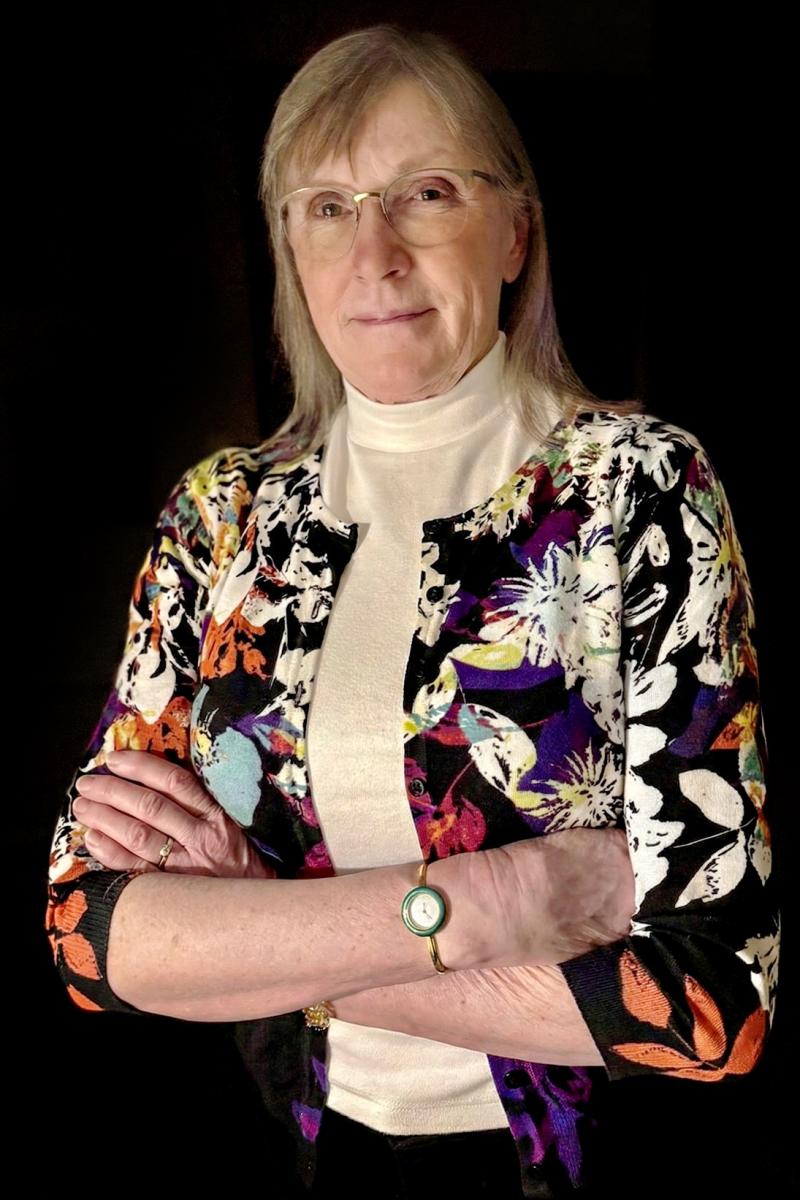 Lynda Cooper Photo en couleur d’une femme d’âge mûr portant des lunettes et une veste à motifs floraux, les bras croisés, Lynda Cooper.