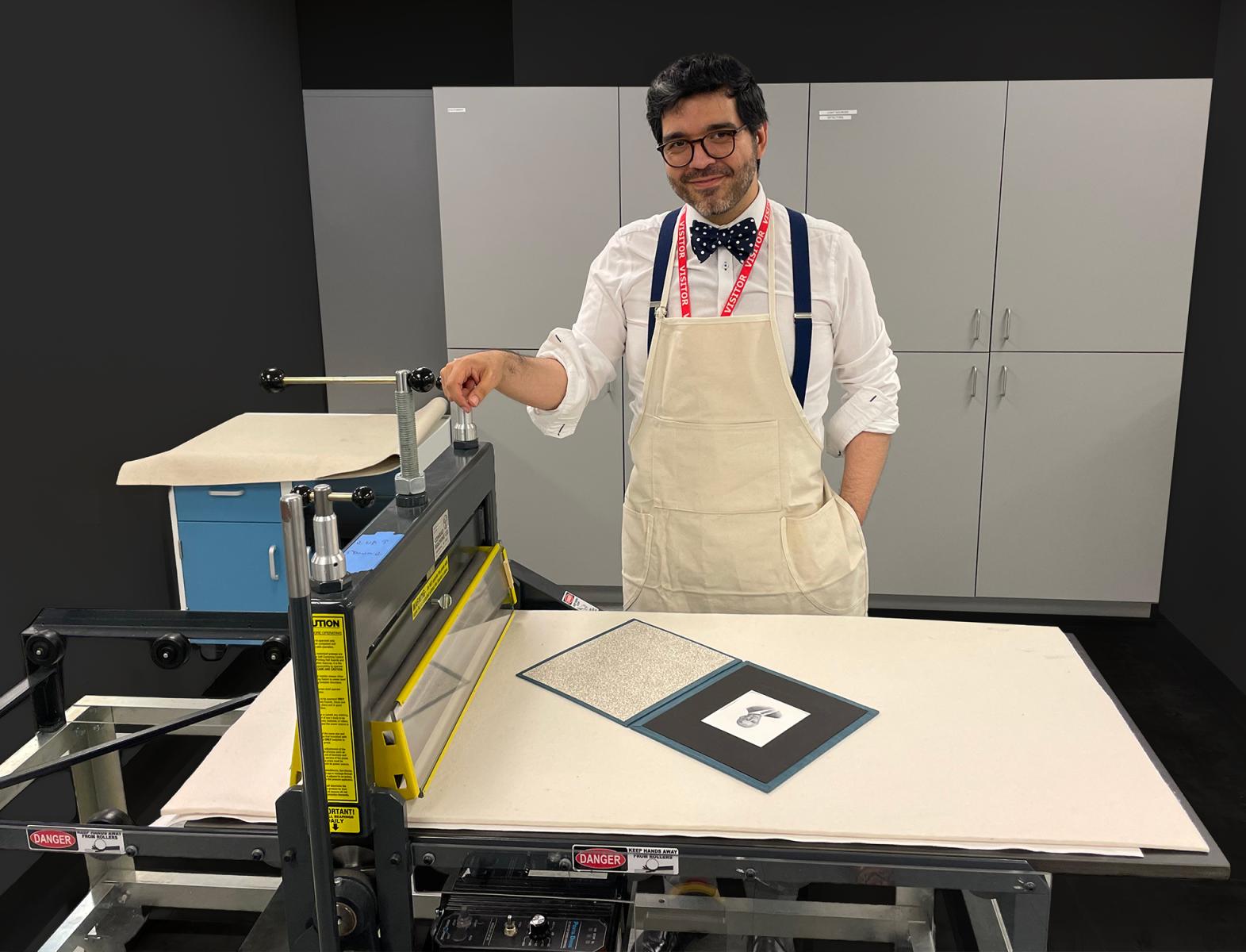 Maître graveur Homme portant un tablier qui se tient debout derrière une presse, dans un atelier, à une table où se trouve une épreuve de gravure.
